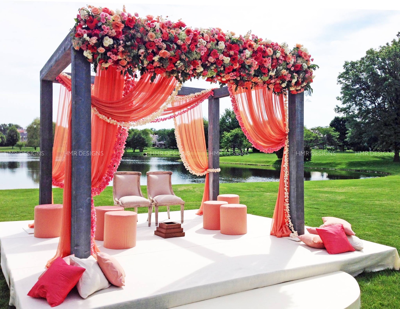 A steel framed canopy or mandap is draped with peach, pink and coral fabric and flowers for an outdoor Indian wedding by HMR Designs